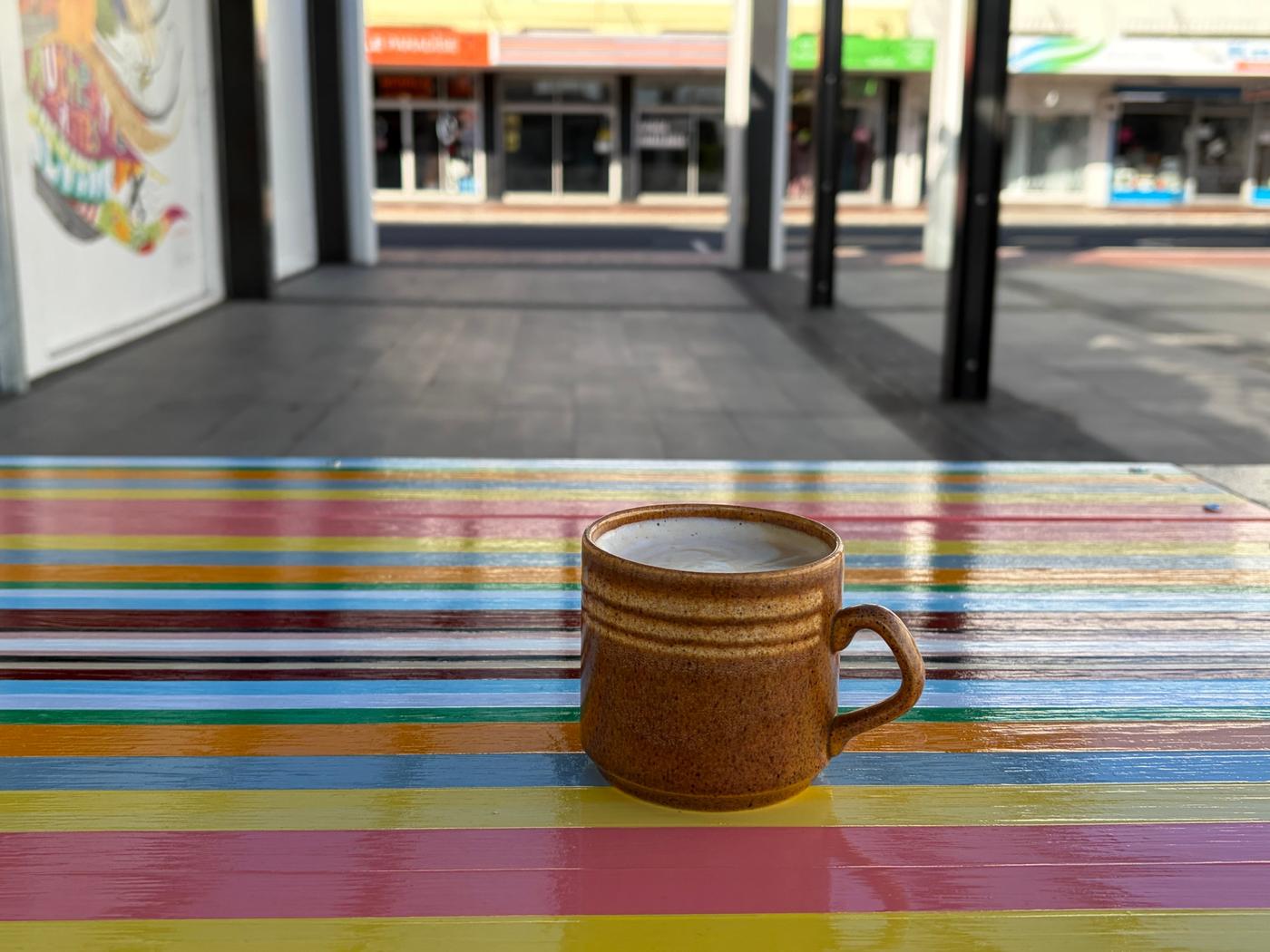 A speckled brown mug of coffee on a glossy table painted in Strip Painting bands, in the 300 West laneway, looking out across Heretaunga Street West to the shops on the south side