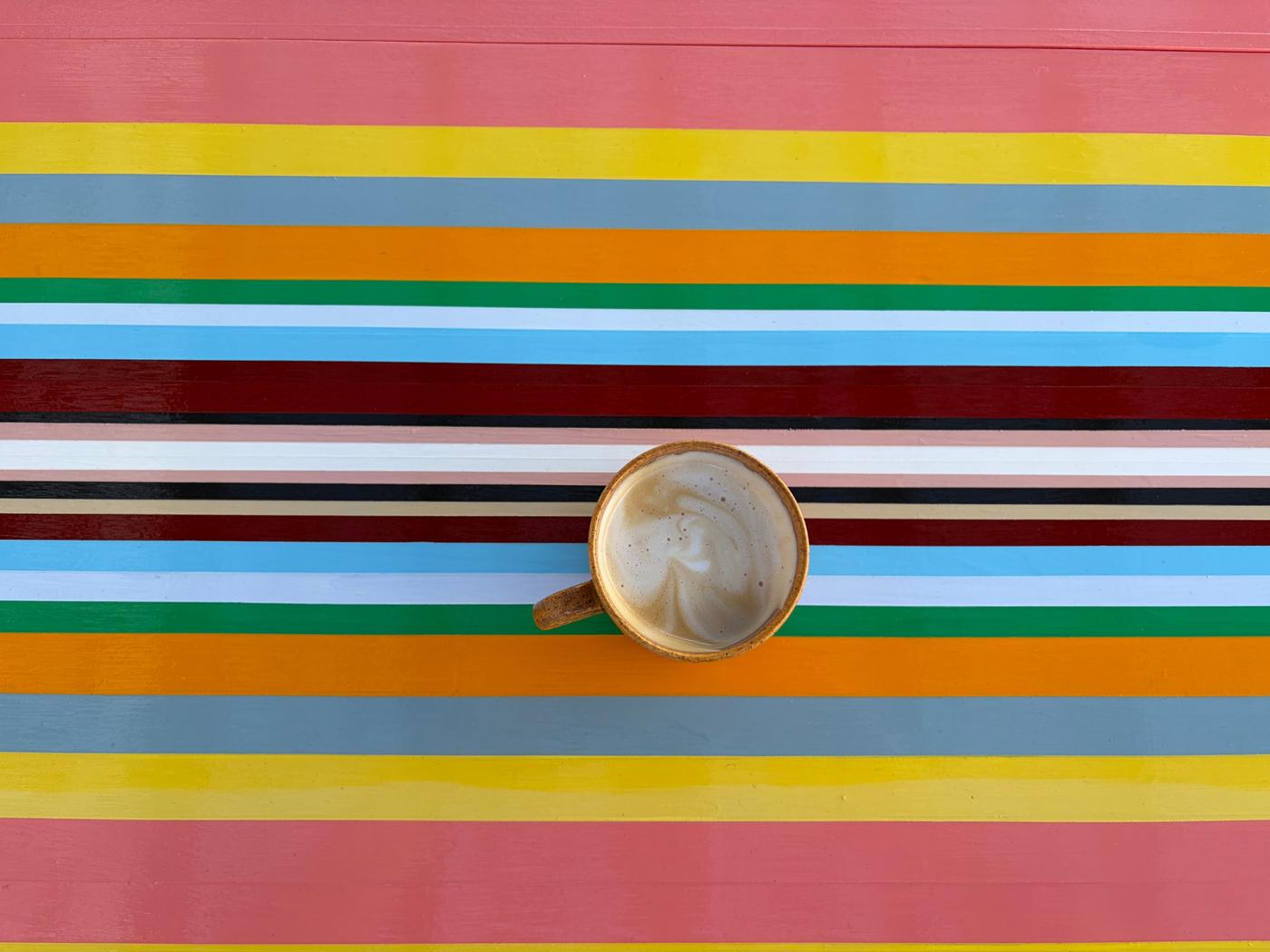 Looking straight down at a coffee in a speckled brown mug on a table painted in Strip Painting bands of coral, yellow, grey, orange, green, sky blue, maroon, blush and black