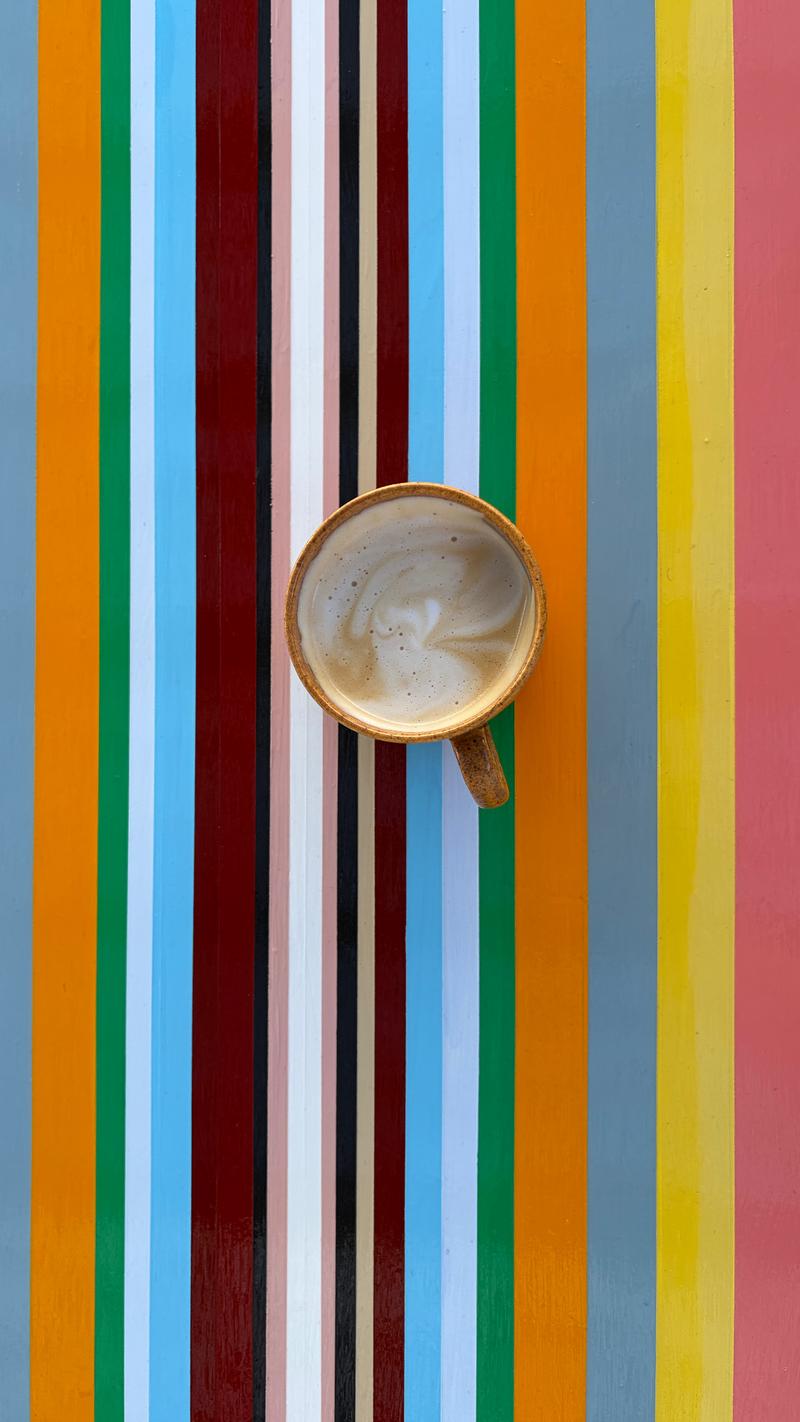 Looking straight down at a coffee on the Strip Painting table, its bands running lengthways in grey, orange, green, sky blue, maroon, black, blush, white, yellow and coral
